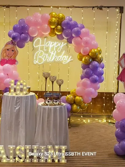 A Barbie themed birthday party is always a hit. This setup features a 'Happy Birthday' neon sign against a fairy light curtain, a pink and purple balloon arch, and Barbie character cutouts to bring the theme to life.