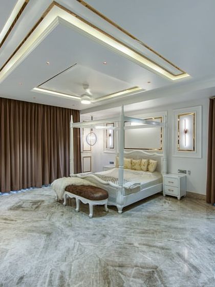 A grand, neoclassical-inspired bedroom with a four-poster bed, ornate ceiling details with gold accents, and classic wall paneling. The expansive marble floor enhances the sense of luxury.