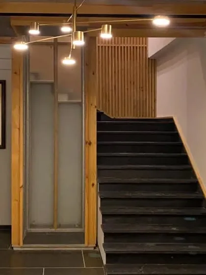 The staircase and elevator area in the Lucknow penthouse, showing the contrast between the dark stone steps, warm wood paneling, and the soft light from a modern brass chandelier.
