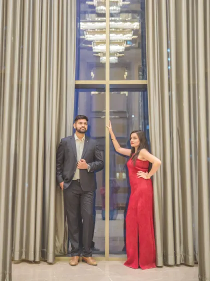 A dramatic, full-length portrait of the couple standing against floor-to-ceiling curtains. The window behind them reflects the city lights, adding a touch of urban glamour.