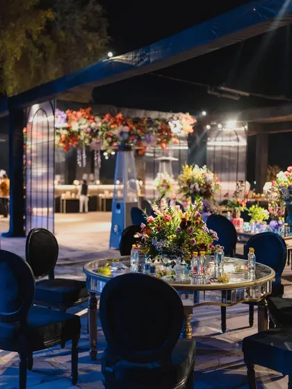 An elegant outdoor Sangeet setup with mirrored tables, black velvet chairs, and towering floral arrangements, combining classic glamour with an open-air setting.