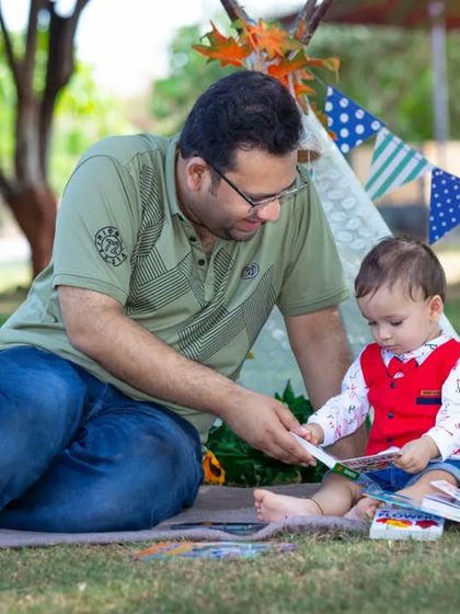 A father and his one-year-old son are engrossed in a picture book during a picnic-style photoshoot, complete with a teepee and colorful toys.