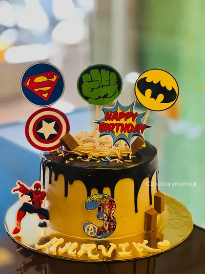 An action-packed superhero cake for a 3rd birthday. It features logos of Superman, Hulk, Batman, and Captain America, with a chocolate drip and an Avengers theme.