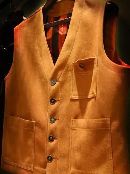 This tan suede vest is a perfect layering piece. The image focuses on its practical pockets and button details, showing how we blend utility with refined style.