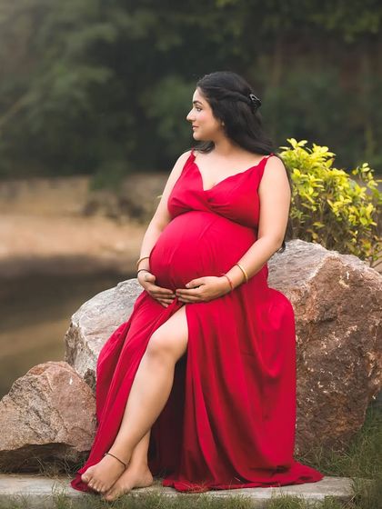 A stunning full-length portrait of a mother-to-be in a vibrant red dress, seated on a rock in a park. The natural green background makes the red color pop, creating a striking image.