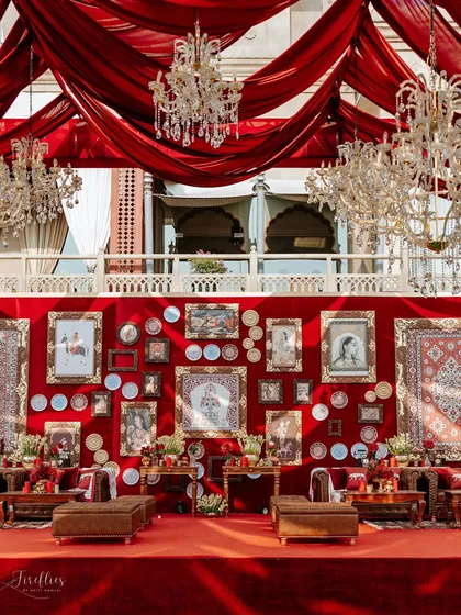 The stunning lounge area for the 'Laal Ishq' wedding, featuring a rich red velvet backdrop adorned with traditional paintings, carpets, and decorative plates. The draped ceiling and chandeliers complete the luxe, heritage look.