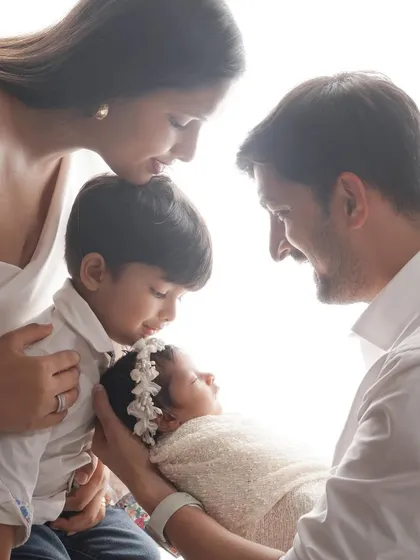 A tender family moment captured with a high-key lighting effect, showing parents and an older brother lovingly looking at the new baby in their arms.