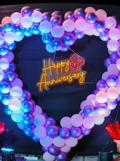 A stunning heart-shaped balloon frame for an anniversary. The frame is made of purple and white balloons and surrounds a 'Happy Anniversary' neon sign, creating a perfect photo booth.