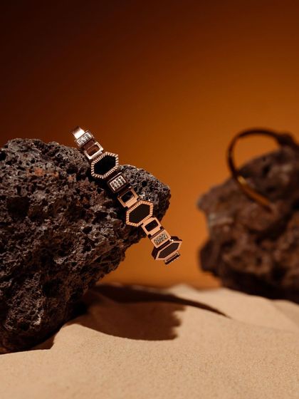 A geometric bracelet resting on a volcanic rock against a sandy background. The composition is earthy, raw, and luxurious.