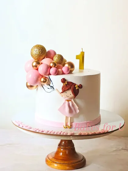 A sweet and simple first birthday cake featuring a cute fondant girl holding a cluster of pink and gold chocolate balloons.