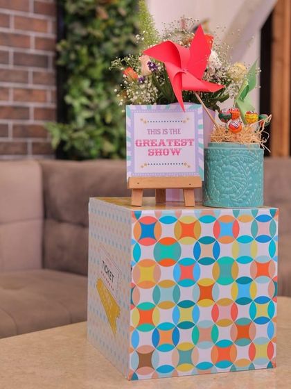 A colorful centerpiece on a geometrically patterned box. The arrangement includes a red pinwheel, fresh flowers, and a mini "This is the Greatest Show" sign, perfect for a carnival-themed table.