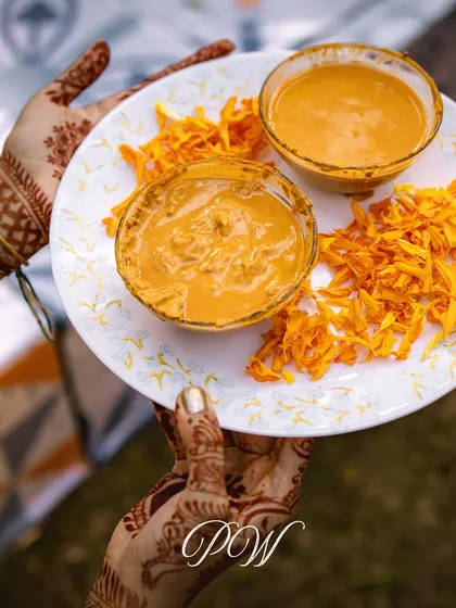 A close-up of a henna-adorned hand holding a plate with Haldi paste and marigold petals, capturing the essence of the ceremony.