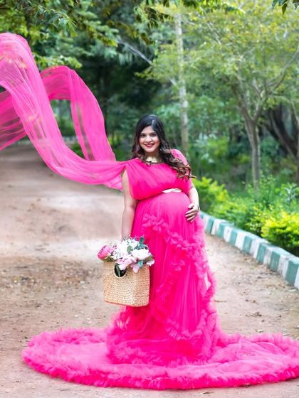 A dynamic and joyful shot of a mom-to-be in a pink gown with a long, flying train. She's walking down a path holding a basket of flowers, looking absolutely radiant.