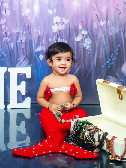 A little girl dressed as a mermaid with a red tail sits with a treasure chest for her first birthday themed photoshoot.