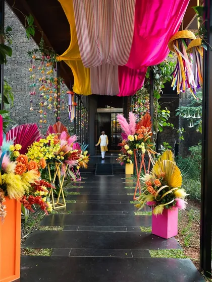 The entrance walkway to the Haldi, lined with vibrant, modern floral arrangements in pink and orange, leading into the main event area.