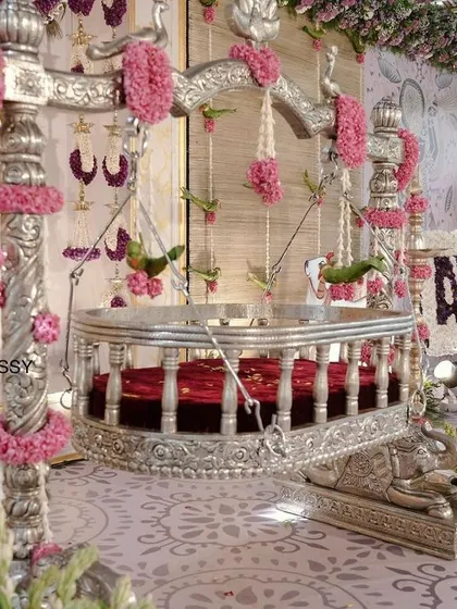 A close-up of the beautifully decorated cradle, adorned with pink floral tassels. The name of the baby, "Avyukth," is displayed on a floral board to the side, making it a central part of the decor.