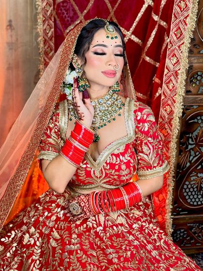 A beautiful bride in a classic red lehenga with green jewelry. The makeup features soft, shimmery eyes and a natural-looking base.