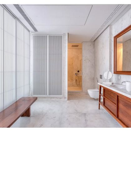 A spacious master bathroom in the Bombay Penthouse. White marble clads the floors and walls, creating a bright and serene sanctuary, while a teak bench and vanity add warmth.