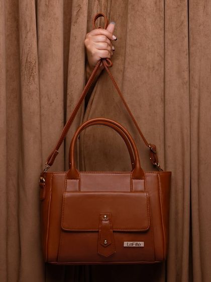 A hand holding a tan handbag against a matching brown curtain. This simple, elegant shot focuses on the product's color and form.