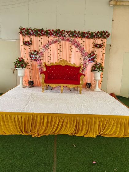 An outdoor wedding stage setup on an artificial grass lawn. The peach drapes and floral arch provide a soft, romantic backdrop against the open-air setting.