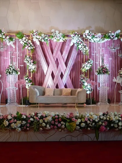 A sophisticated reception stage with a backdrop of woven pink drapes, framed by an arch of white and pink flowers. The classic white urns add a touch of timeless elegance.