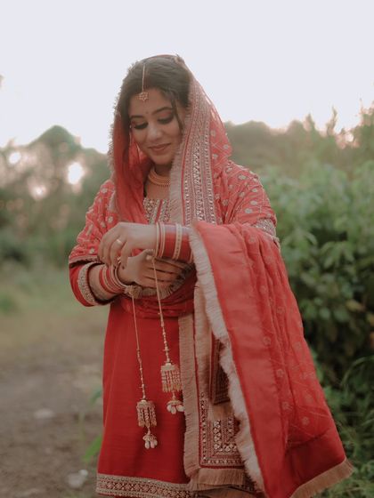 A beautiful solo shot of the bride in her traditional red suit, adjusting her kaleere. This image focuses on the intricate details of bridal attire and quiet, personal moments.