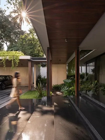 The arrival experience at Villa Shakthi is defined by a covered veranda and driveway flanked by lush greenery. The play of light and shadow under the wooden ceiling, combined with the reflection on the polished granite floor, creates a welcoming and dynamic entrance.