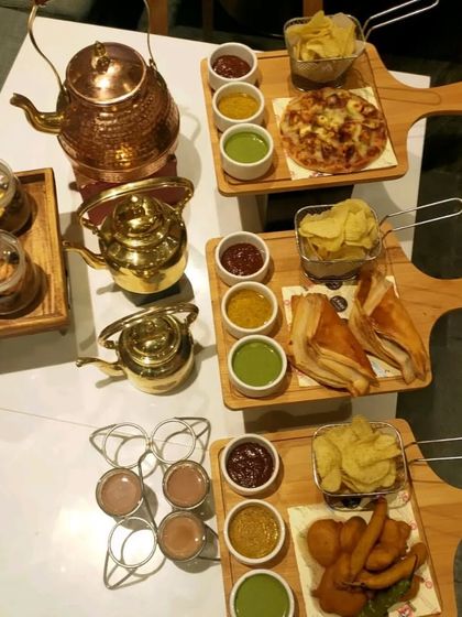 A perfect spread for a high-tea event. This includes a variety of snacks like sandwiches, pakoras, and mini pizzas, served with different chutneys and chai in traditional kettles.