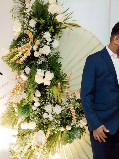 An elegant engagement backdrop for Rhea and Jeevan. Their initials are prominently featured amidst a beautiful arrangement of white florals and green foliage, creating a perfect photo corner.
