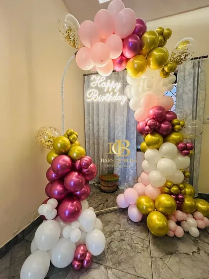 A bright and cheerful birthday decoration in shades of pink, magenta, and gold. This setup features a balloon arch over a simple curtained backdrop, with a neon sign adding a warm glow.