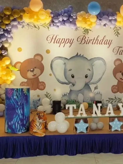 A cute and classic animal-themed first birthday stage. This setup features an elephant and teddy bear backdrop, a colorful balloon arch, and marquee letters spelling out the birthday boy's name.