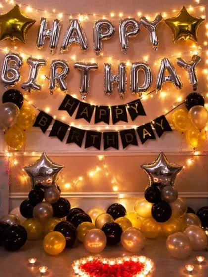 A beautiful and cozy birthday setup in a room. This decoration features a wall of fairy lights, "Happy Birthday" foil balloons, and clusters of black, gold, and silver balloons on the floor.