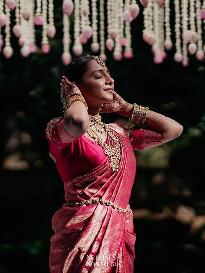 A bride in a pink saree, her face tilted towards the sun, stands under a canopy of hanging lotus buds.