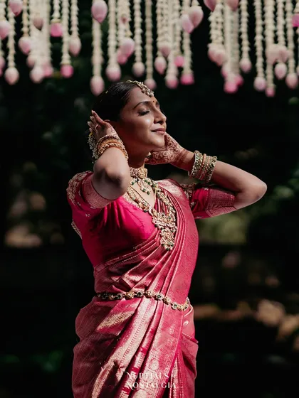 A bride in a pink saree, her face tilted towards the sun, stands under a canopy of hanging lotus buds.