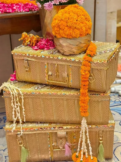 Decorative props for a Garba celebration, featuring stacked wicker trunks with sequin and gota patti borders. A marigold garland and a small golden elephant figurine add a touch of traditional charm.