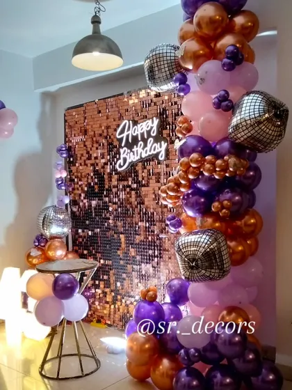 A stunning rose gold shimmer wall creates the perfect photo backdrop, complemented by a custom 'Happy Birthday' neon sign, a cascade of metallic and purple balloons, and sparkling disco balls for a truly glamorous party vibe.