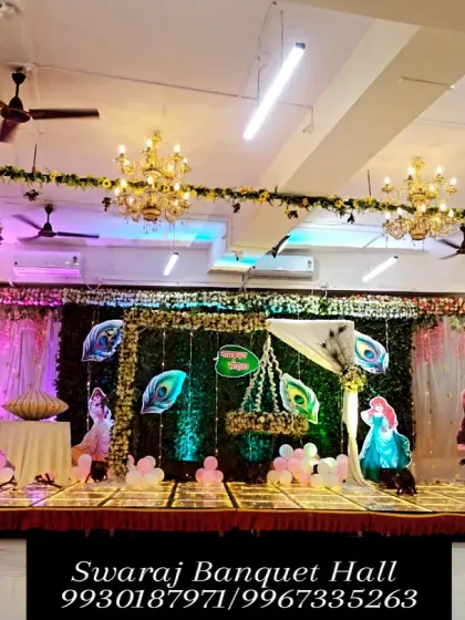 A wide view of a naming ceremony setup in our hall. The stage is decorated with a green turf backdrop, peacock feather elements, and a traditional cradle, with ample seating arranged for guests.