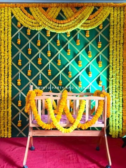 A simple and traditional naming ceremony decoration for home. The setup features a marigold-decorated cradle against a green backdrop with a lattice pattern.