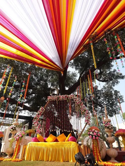A burst of color for a joyous Haldi ceremony! We used vibrant yellow, pink, and orange drapes with traditional marigold flower hangings to create a festive outdoor setup under a beautiful old tree.