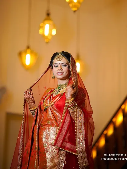 A portrait of a North Indian bride in a vibrant red saree, her veil delicately held. The warm indoor lighting creates a classic, timeless feel.