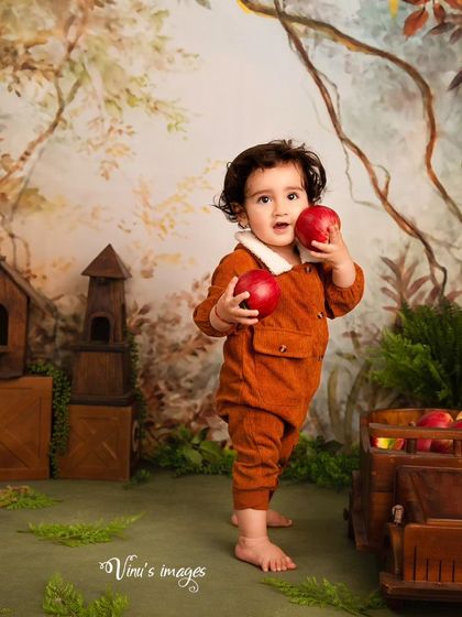 An apple a day. This toddler portrait captures a moment of discovery as the child holds up two bright red apples in our charming, hand-painted forest backdrop.