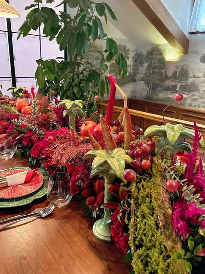 This long view of the tablescape highlights its scale and richness. The runner overflows with red flowers, fruits, and greenery, creating a continuous line of color and texture down the length of the dining table.
