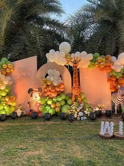 An outdoor "Wild One" setup with a beautiful balloon arrangement in earthy tones of green and orange, complete with zebra and giraffe props against the natural backdrop of palm trees.