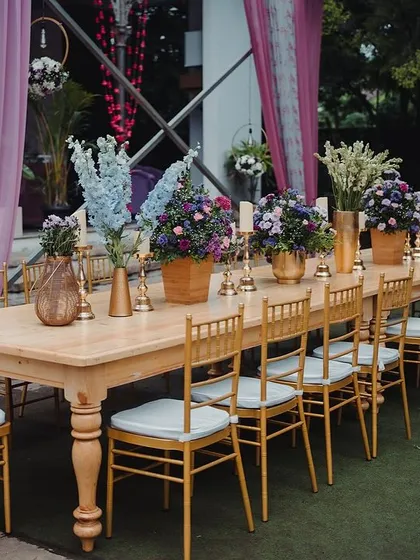 A long wooden table set for a reception dinner, with beautiful floral centerpieces and golden chairs.