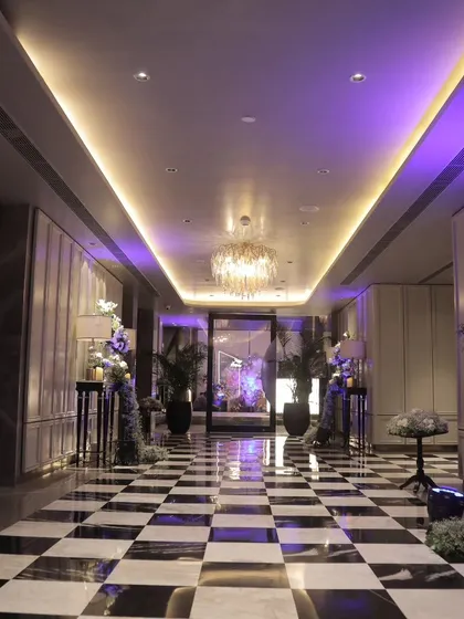A chic and modern hotel corridor transformed into a grand entrance. The checkerboard floor, purple uplighting, and floral arrangements create a sophisticated and stylish pathway for guests.