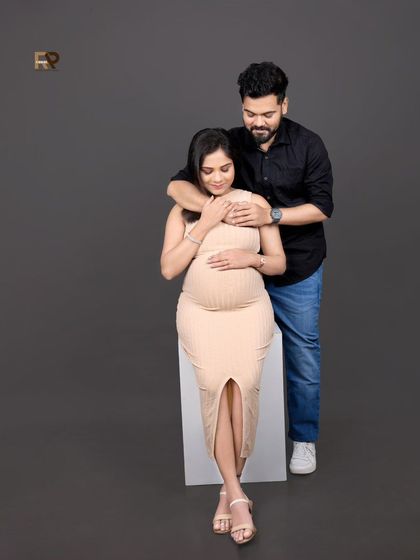 A stylish and modern maternity portrait. The couple's coordinated neutral and black outfits create a chic, minimalist aesthetic against the solid grey background.