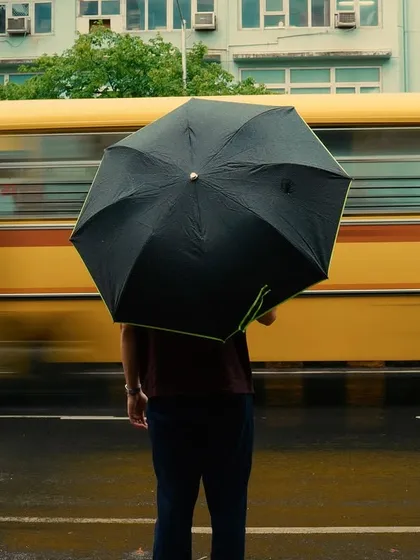 A person stands with an umbrella, perfectly aligned with a yellow school bus blurring past. The color coordination and motion create a visually striking and well-timed street photograph.