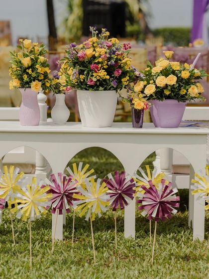 Charming details from the pastel Haldi decor. A row of hand-painted pinwheels stands in front of a white bench adorned with fresh yellow and purple flowers in pastel vases.