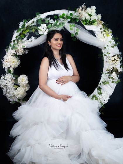 Ethereal beauty. This mom-to-be looks serene and angelic in a white gown, seated in our floral hoop prop against a simple black background.