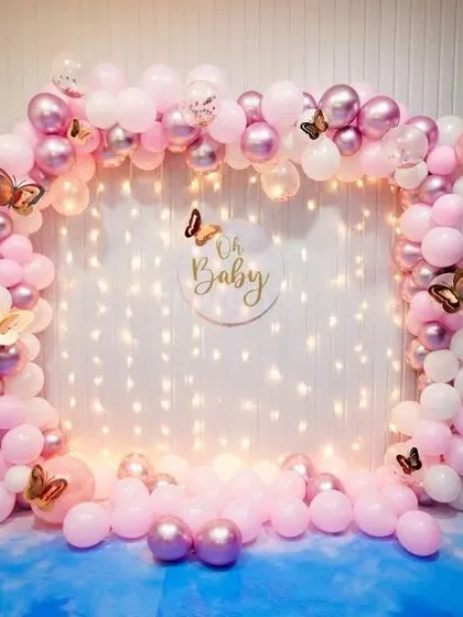 A delicate and pretty "Oh Baby" backdrop for a baby shower. The pink and white balloon arch is decorated with butterfly accents and set against a wall of fairy lights.